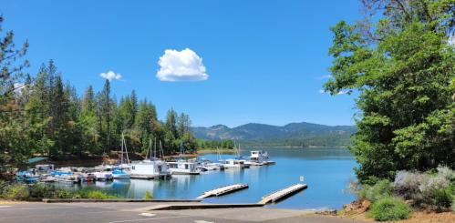 A serene lake scene with boats docked, surrounded by trees and mountains under a clear blue sky.